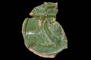 Fragment des Innenfelds einer Halbzylinderkachel mit geschlossenem Vorsatzblatt mit Ritter beim Gestech in rundem Medaillon mit losem Tauband, grün glasiert, um 1450, Emmendingen, Hochburg